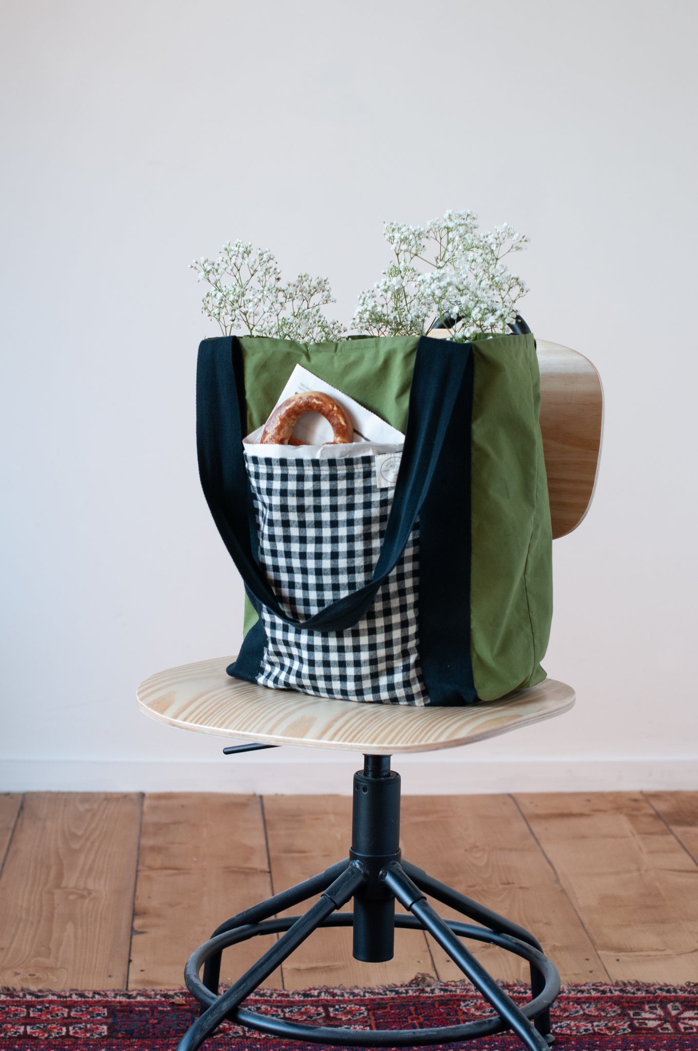 A green and black checkered tote bag with a plaid pattern, featuring two large side pockets and a smaller interior pocket, displayed on a wooden stand with a white flower arrangement in it.