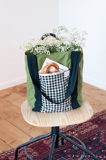A green and black checkered tote bag with a plaid pattern, featuring two large side pockets and a smaller interior pocket, displayed on a wooden stand with a white flower arrangement in it.