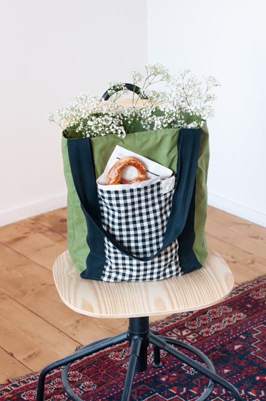 A green and black checkered tote bag with a plaid pattern, featuring two large side pockets and a smaller interior pocket, displayed on a wooden stand with a white flower arrangement in it.