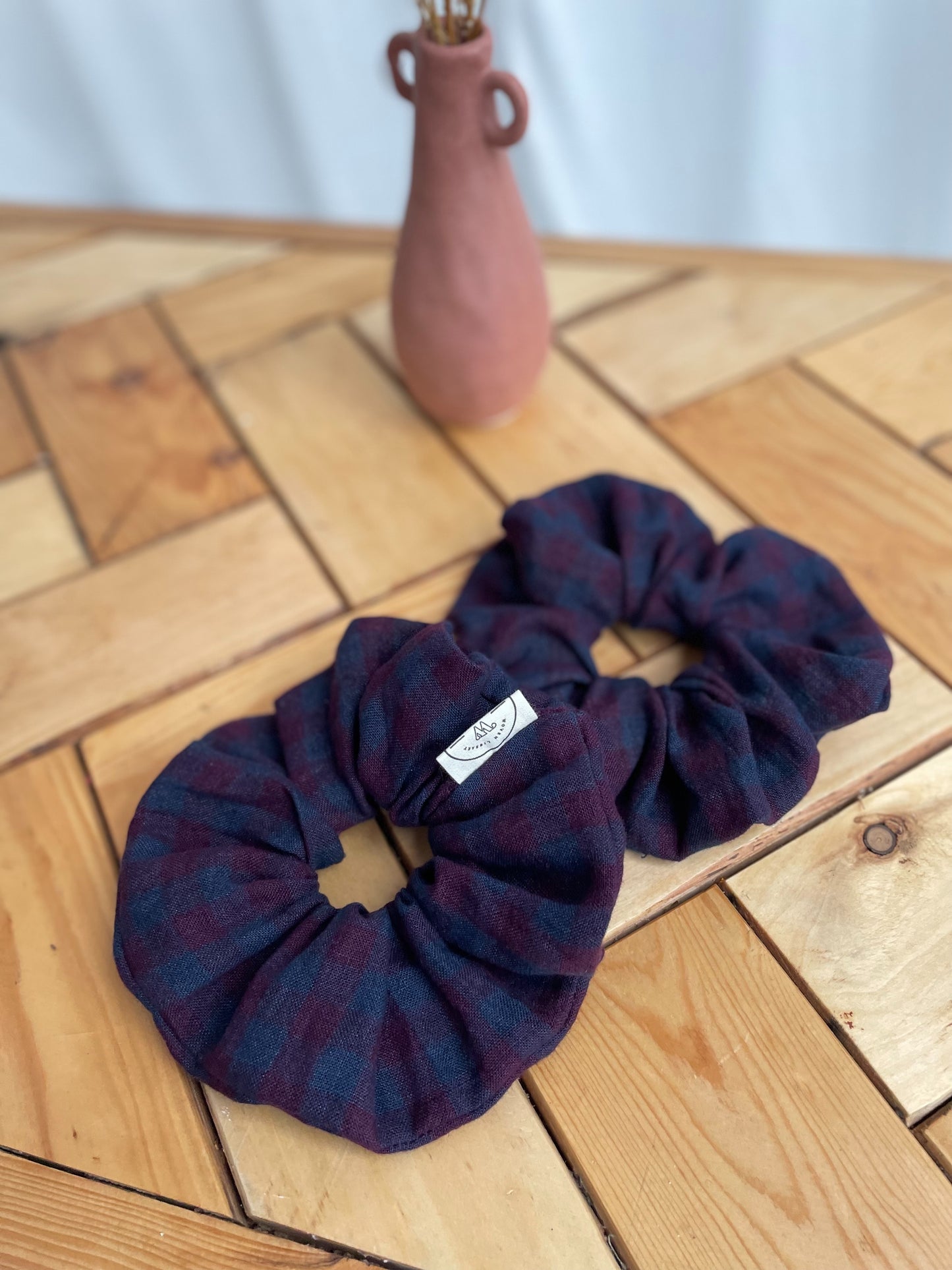 Two purple linen checked scrunchies on a wooden surface with a vase in the background.