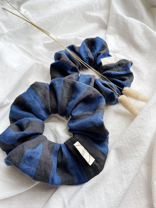 Two striped scrunchies in black and blue, one with a label attached, displayed on a white textured surface.