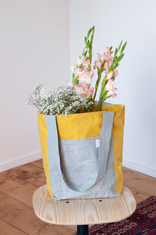 A yellow cotton tote bag with a grid of exterior pockets, filled with flowers, placed on a wooden surface.