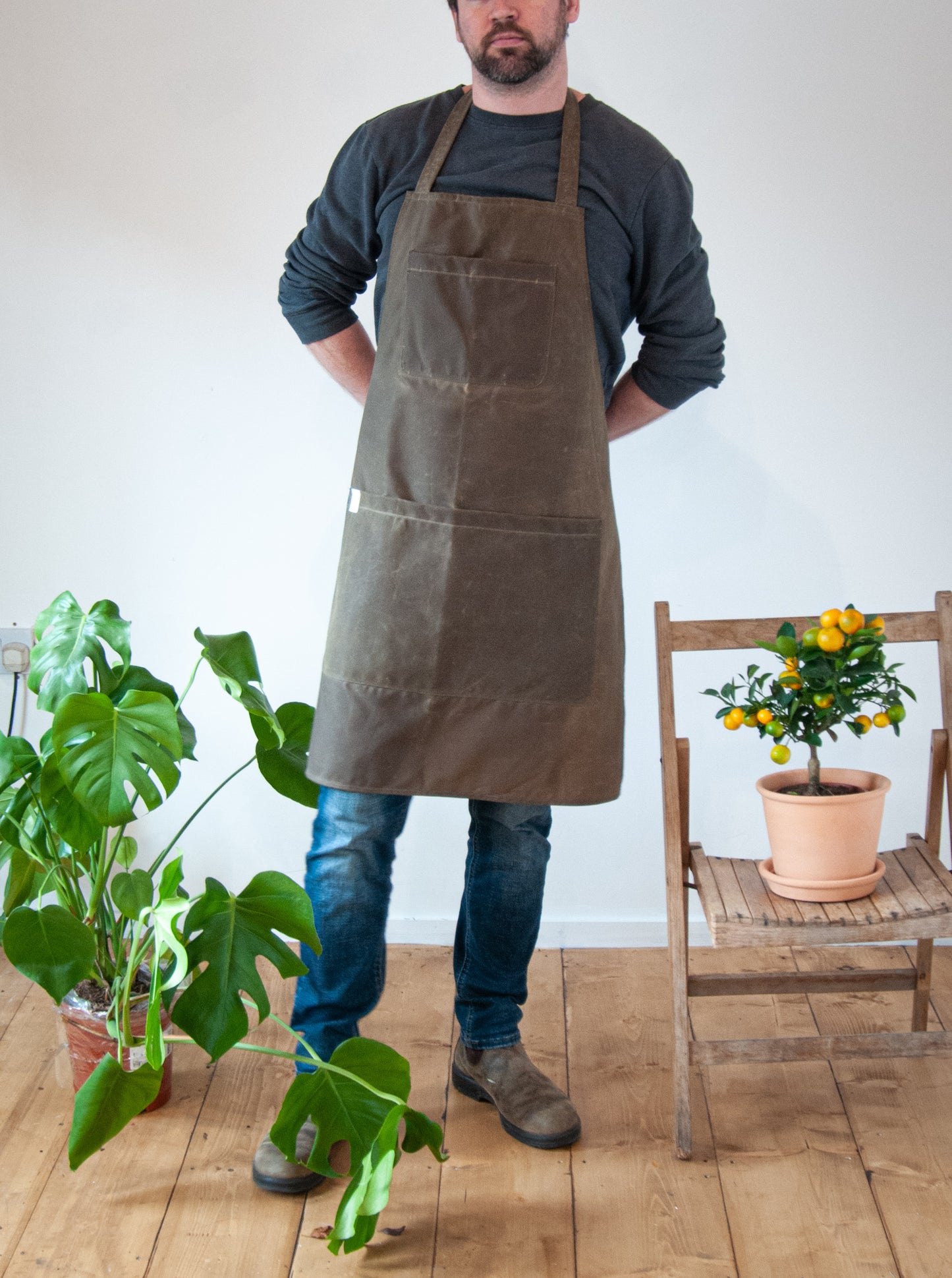 A person wearing a khaki waxed cotton apron with pockets, standing in a room with potted plants and a wooden floor.
