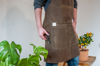 a man wears a wax cotton apron in a brown khaki colour beside some plants
