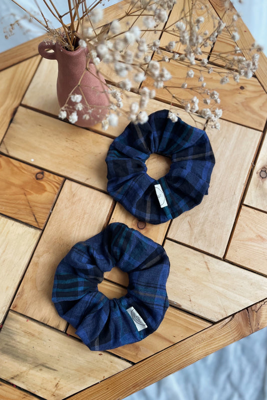 Two blue scrunchies with a plaid design on a wooden surface with a vase of dried flowers in the background.