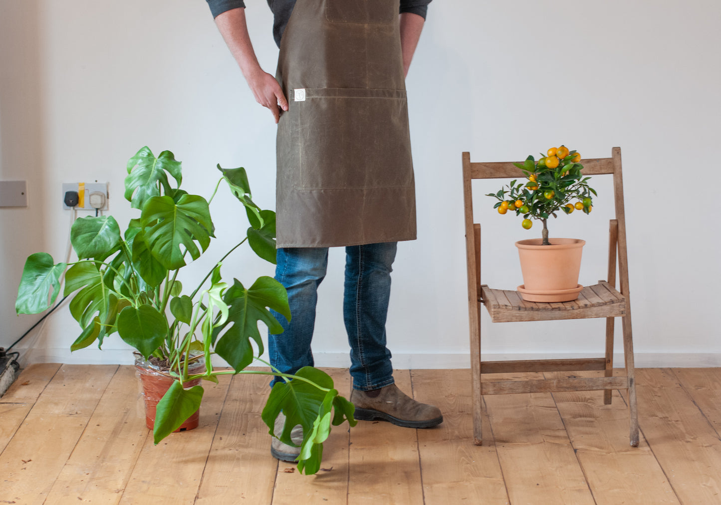 a man wears a brown wax cotton apron beside some plants and standing on a wooden floor